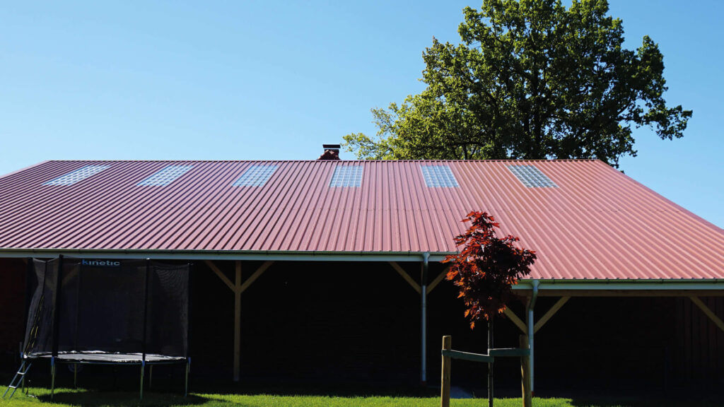 Dak met rode trapeziumplaten, lichte panelen en omringd door groene bomen.